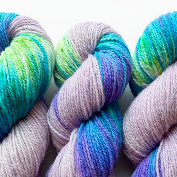 Three skeins of variegated yarn with pastel lavender, teal, green, and purple tones, arranged closely together on a white background.