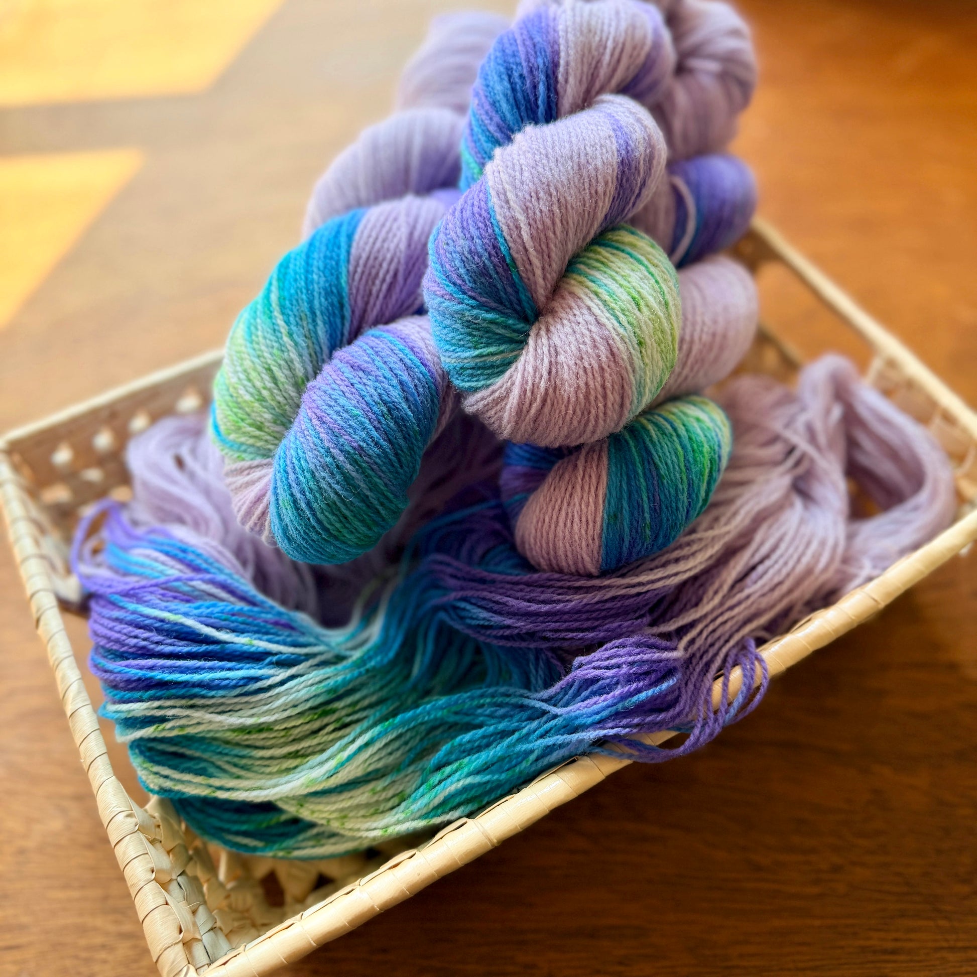 Three skeins of variegated yarn in pastel shades of purple, blue, green, and lavender, arranged in a woven basket on a wooden table.