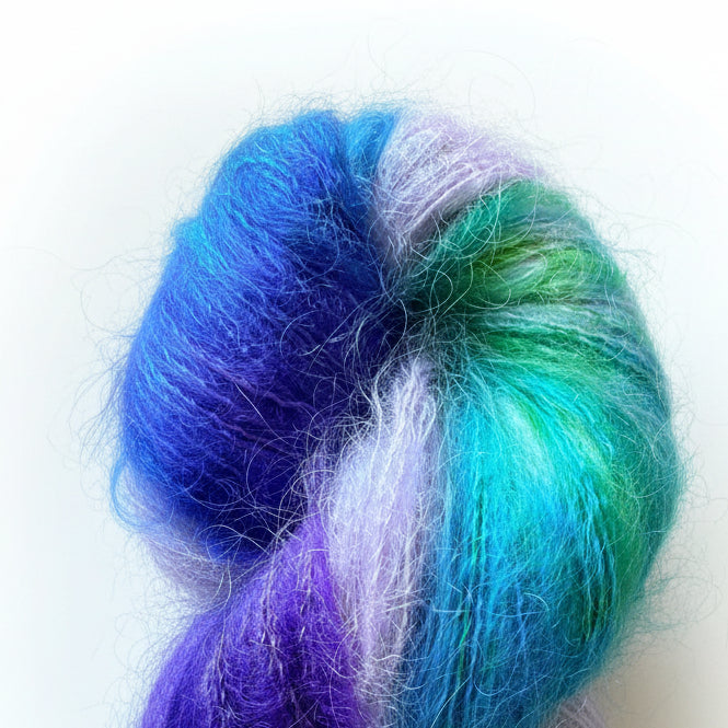 A skein of fluffy variegated yarn resting on a wooden surface. The yarn features blended strands of bright blue, purple, green, and pale lavender.