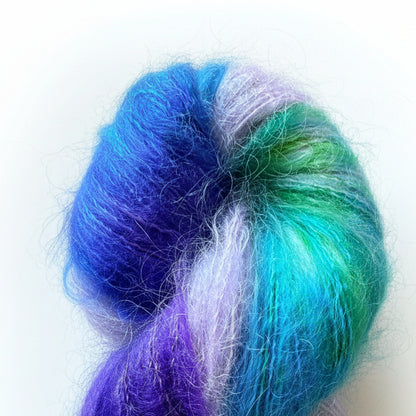 A skein of fluffy variegated yarn resting on a wooden surface. The yarn features blended strands of bright blue, purple, green, and pale lavender.