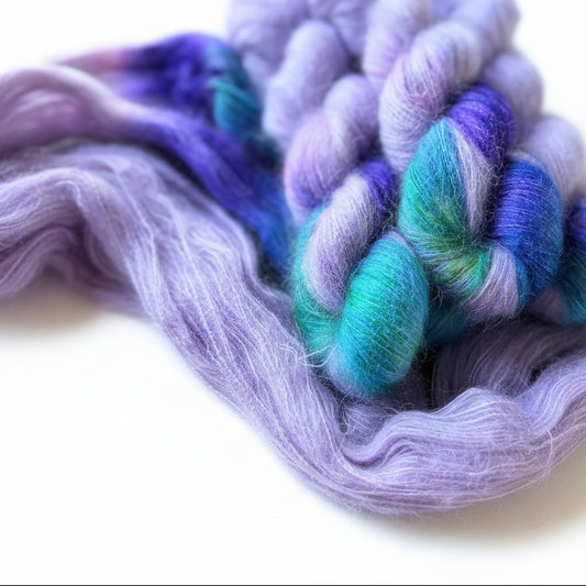 A close-up of variegated wool roving and a twisted yarn skein on a wooden surface, showing blended lavender, purple, blue, and teal fibers.
