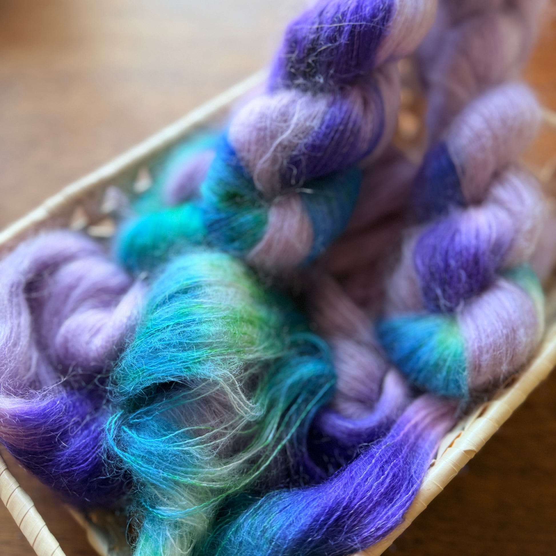 Three skeins of soft, fuzzy yarn in a woven basket, featuring a variegated color pattern of purple, teal, and lavender on a warm wooden background.