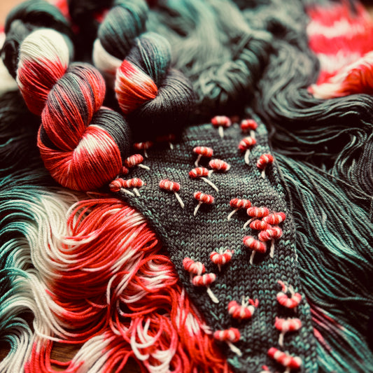 A close-up of a handwoven textile featuring a black base with red berry-like embroidered details and surrounded by variegated yarns in red, green, and cream.