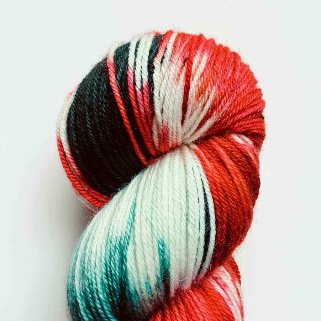A close-up of a twisted skein of variegated yarn on a wooden surface. The yarn features variegated stripes of red, orange, mint green, black, and white.