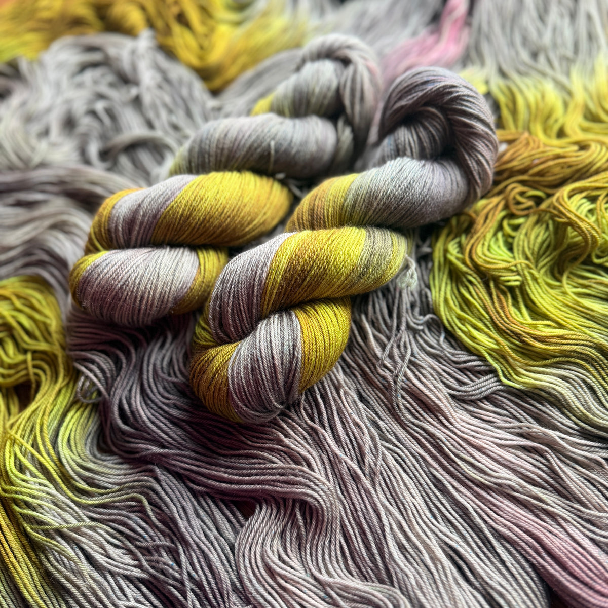 Close-up of assigned pooling yarn skeins in a gradient of yellow, gray, and dusty pink, arranged on a matching pile of yarn.