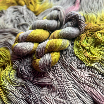 Close-up of assigned pooling yarn skeins in a gradient of yellow, gray, and dusty pink, arranged on a matching pile of yarn.
