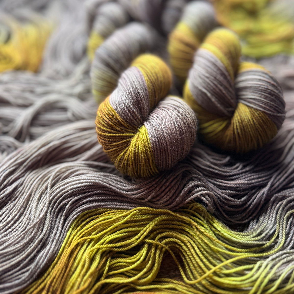 Close-up of several skeins of variegated yarn in gray, taupe, and mustard yellow, arranged on a bed of matching yarn strands.