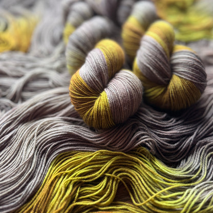 Close-up of several skeins of variegated yarn in gray, taupe, and mustard yellow, arranged on a bed of matching yarn strands.