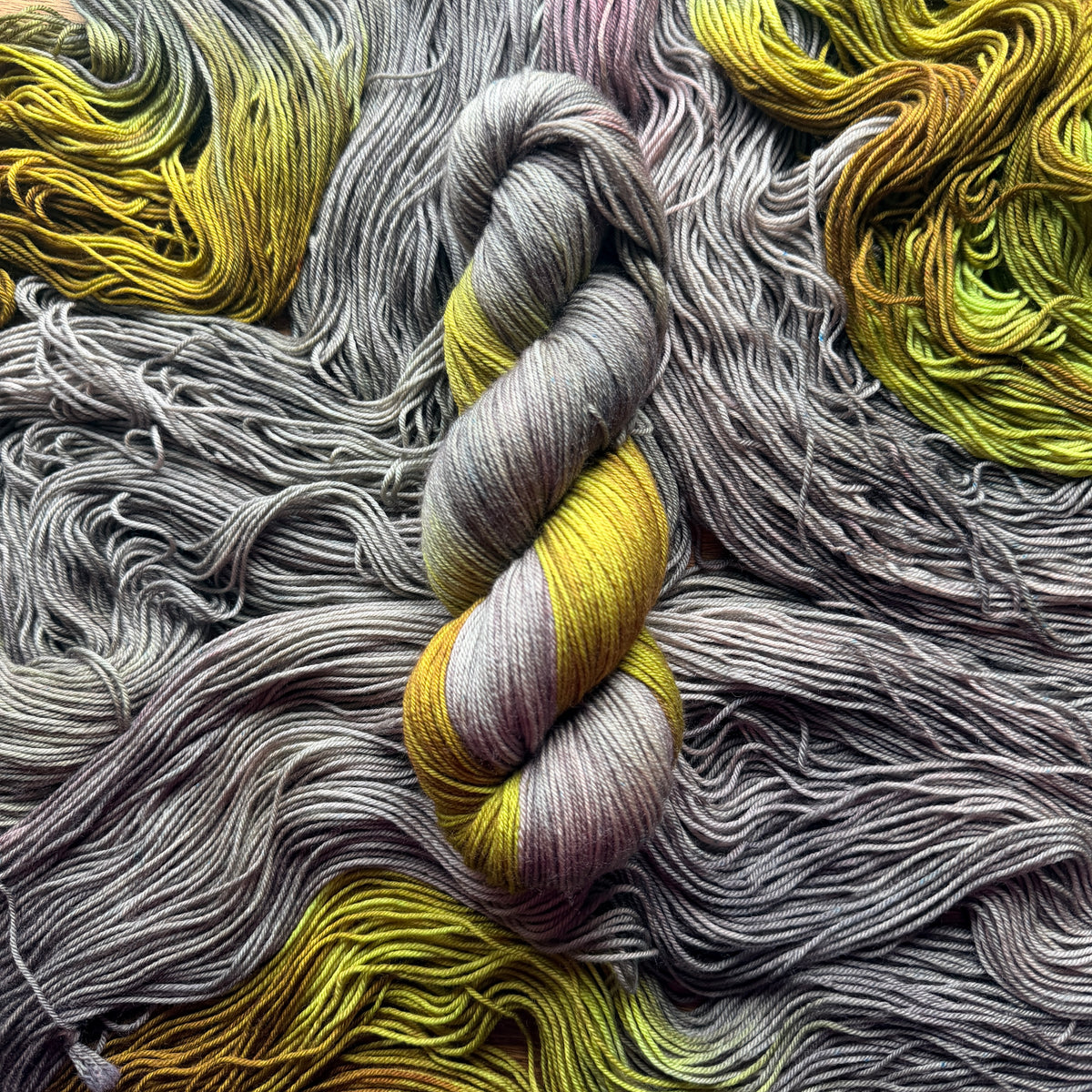 A close-up of variegated yarn arranged in a spiral, with a central twisted skein showing gray, lavender, and mustard yellow tones. 