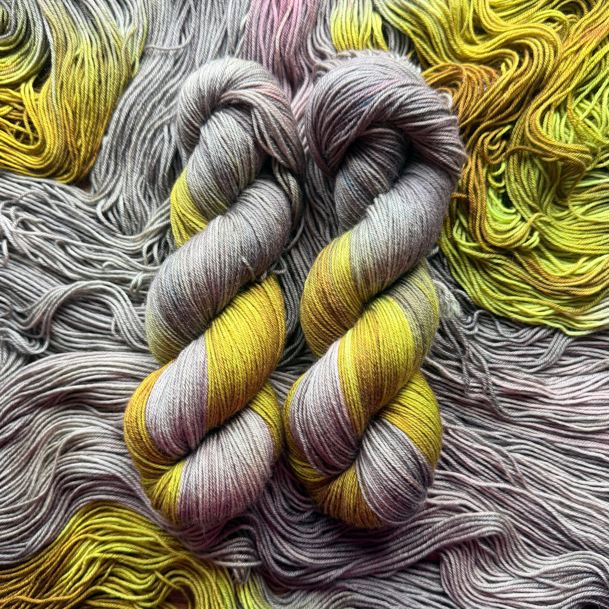 Two twisted hanks of variegated yarn laid flat on a bed of matching yarn. The yarn shows a color-shifting or variegated pattern with yellow, lavender-purple, and gray tones.