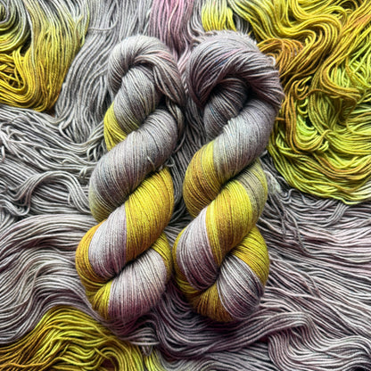 Two twisted hanks of variegated yarn laid flat on a bed of matching yarn. The yarn shows a color-shifting or variegated pattern with yellow, lavender-purple, and gray tones.