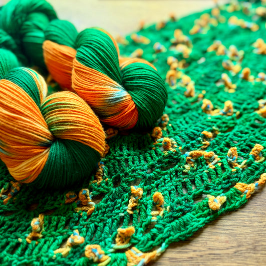Three skeins of variegated yarn in green and orange placed on a green knitted fabric with small orange yarn accents, photographed on a wooden surface.